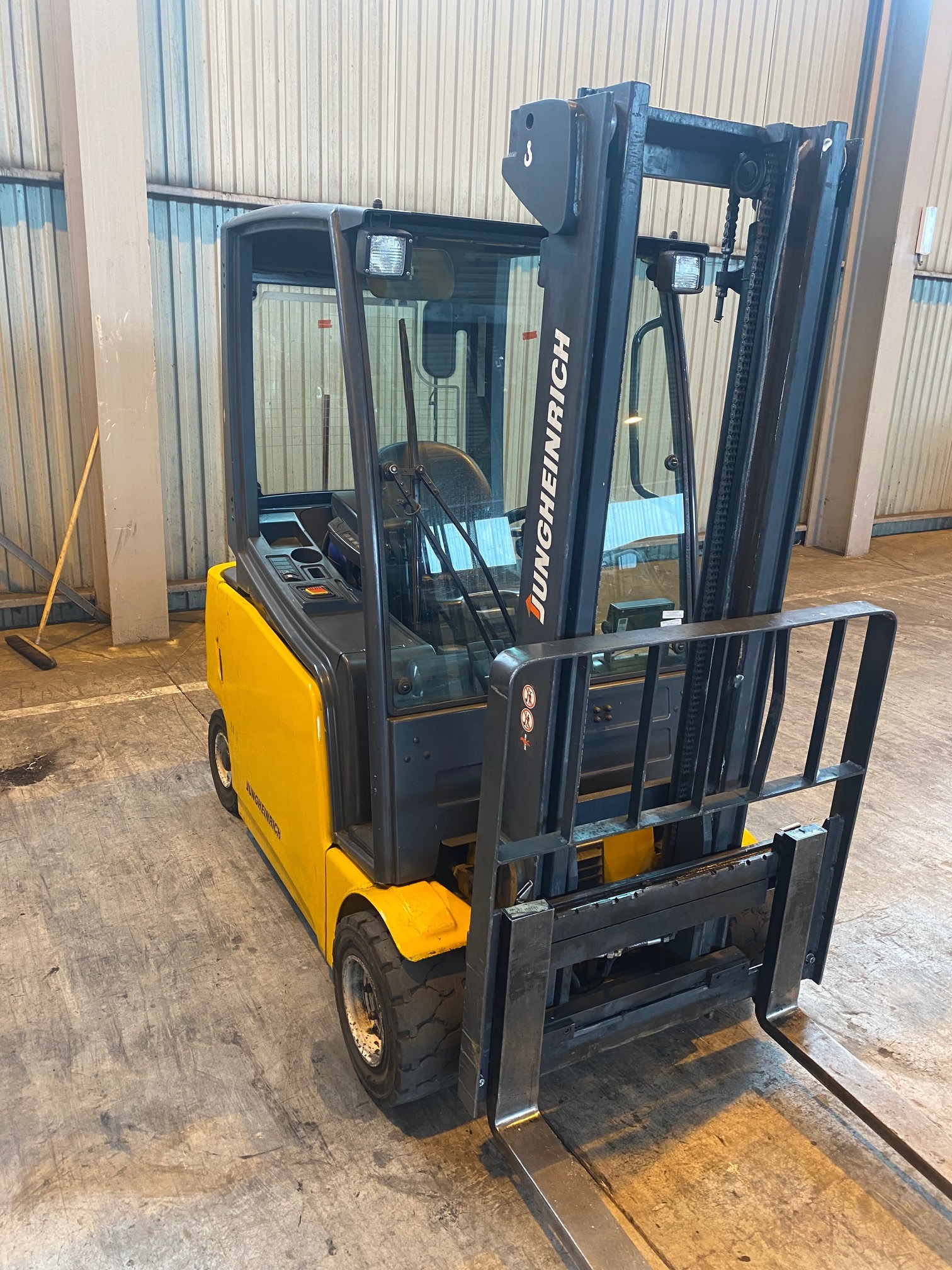 Żółto-czarny wózek widłowy Jungheinrich z kabiną, stojący w hali magazynowej, widok od tyłu.