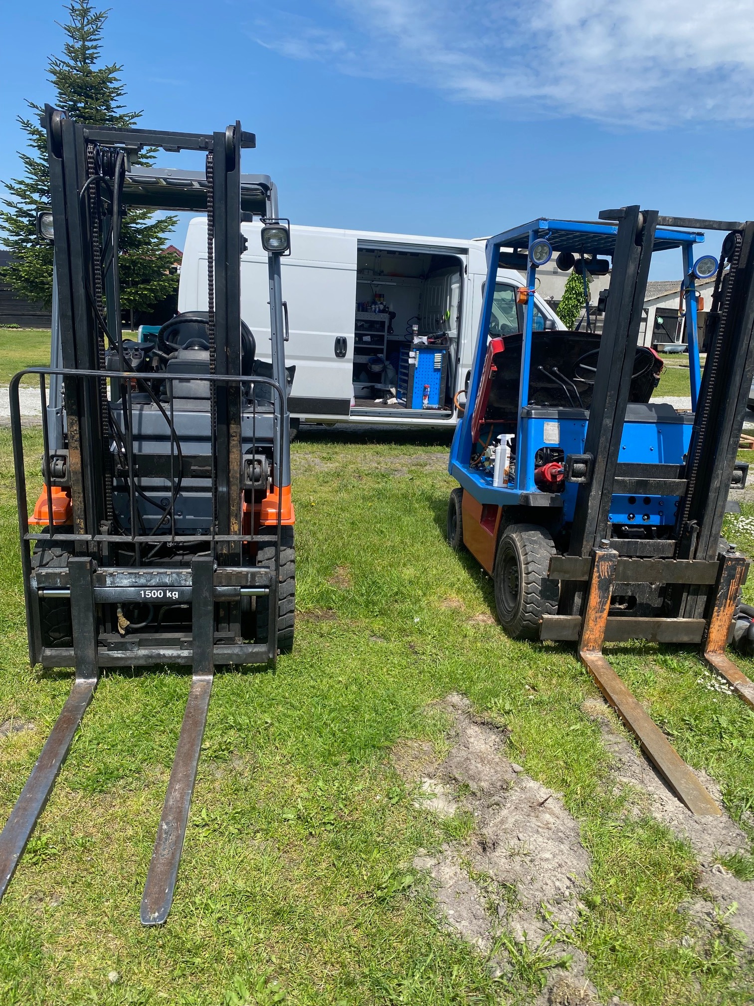 Dwa wózki widłowe (szary i niebieski) stoją obok siebie na trawie, z otwartym vanem serwisowym w tle, sugerującym serwis w terenie.