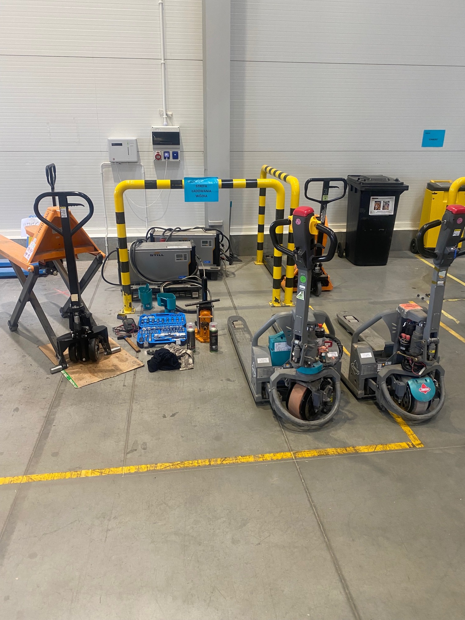 Warsztatowy widok na rozłożone narzędzia i wózki paletowe elektryczne w trakcie naprawy, z widocznymi elementami diagnostycznymi i częściami zamiennymi.