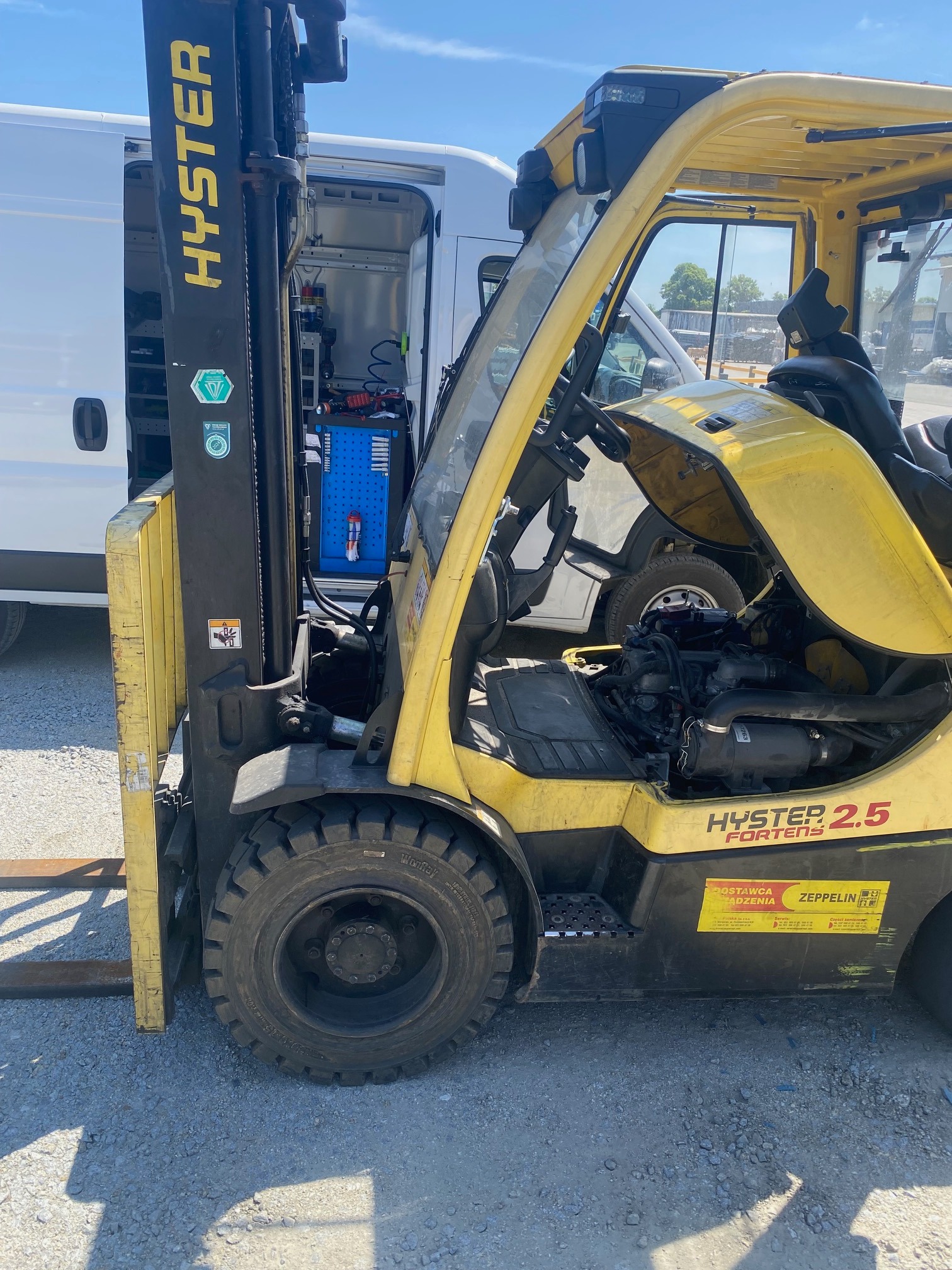 Żółty wózek widłowy Hyster 2.5 z otwartą maską silnika, stojący na żwirowym podłożu, z widocznym wnętrzem furgonetki serwisowej w tle.