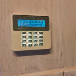 Centrala alarmowa Satel Perfecta z podświetlonym ekranem, wyświetlającym opcje zmiany hasła i użytkowników, zamontowana na pomalowanej ścianie.