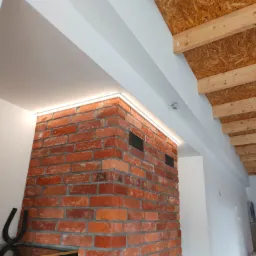 Ceglana ściana z podświetleniem LED w narożniku, widoczna drewniana konstrukcja stropu i fragment pokoju dziecięcego w tle.