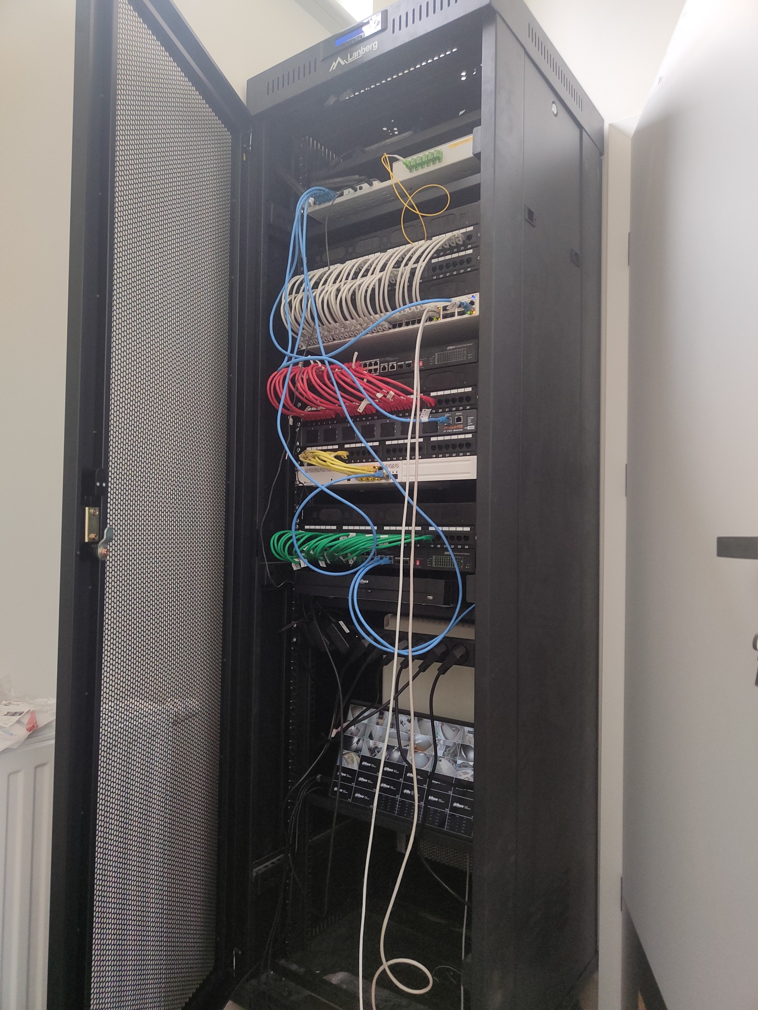 Otwarta szafa rack z zainstalowanymi urządzeniami sieciowymi i okablowaniem w kolorach: niebieskim, białym, czerwonym, żółtym i zielonym. Widoczny switch, router, patch panel oraz rejestrator...