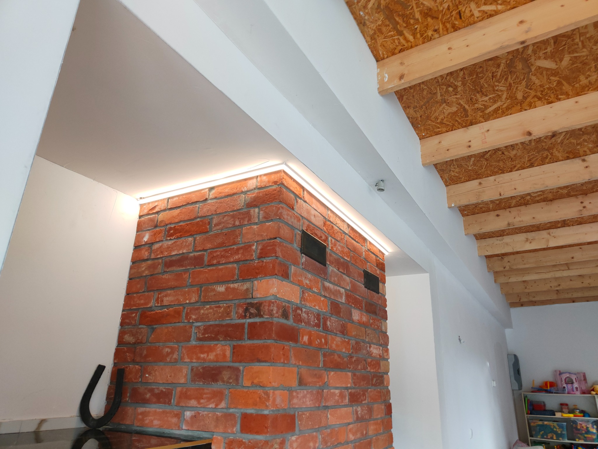 Ceglana ściana z podświetleniem LED w narożniku, widoczna drewniana konstrukcja stropu i fragment pokoju dziecięcego w tle.