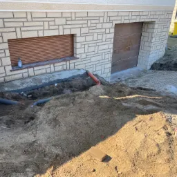 Wykop pod fundamenty domu z widocznymi rurami kanalizacyjnymi i drenażowymi, fragment ściany z okiennicą i butelką płynu do dezynfekcji na parapecie.