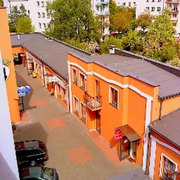 Widok z góry na pomarańczowy budynek z białymi obramowaniami okien i balkonem, otoczony innymi budynkami i zielenią, dwa samochody zaparkowane na dziedzińcu.