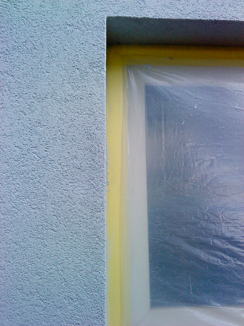 Fragment ściany zewnętrznej z szorstką strukturą, zabezpieczone okno folią i taśmą malarską, przygotowane do prac elewacyjnych.