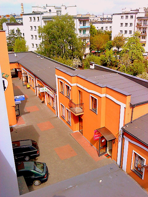 Widok z góry na pomarańczowy budynek z białymi obramowaniami okien i balkonem, otoczony innymi budynkami i zielenią, dwa samochody zaparkowane na dziedzińcu.