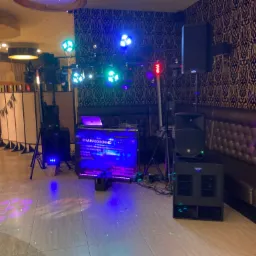 Sprzęt nagłośnieniowy i oświetleniowy ustawiony w sali z dekoracyjną tapetą, widoczne kolumny głośnikowe, mikser DJ-ski z laptopem, reflektory punktowe i ruchome głowy, parkiet oświetlony...