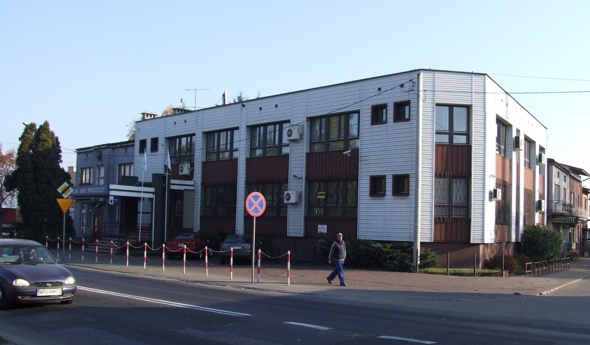 Dwupiętrowy budynek Banku Spółdzielczego przed modernizacją, z widocznymi klimatyzatorami na elewacji i znakiem zakazu parkowania przed wejściem.