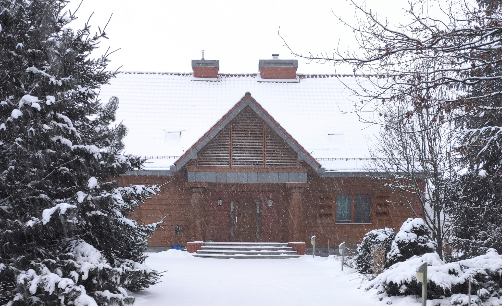 Dworek jednorodzinny z elewacją z desek i cegły klinkierowej, widoczny zimą podczas opadów śniegu, z trzema stopniami wejściowymi pokrytymi śniegiem.