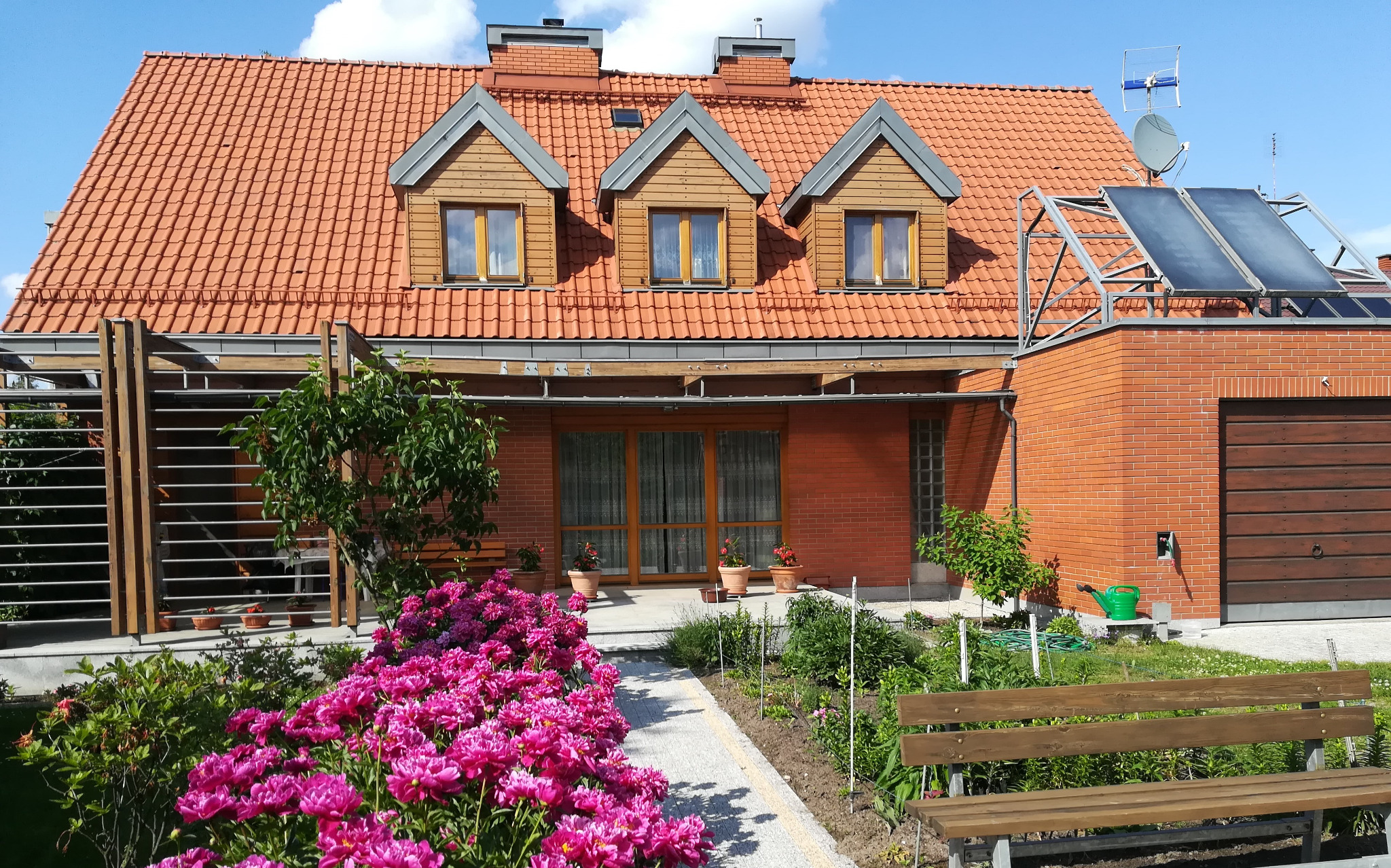 Dwukondygnacyjny dom jednorodzinny z czerwonej cegły i dachówką, z trzema lukarnami i panelami słonecznymi na dachu, otoczony zadbanym ogrodem z kwitnącymi piwoniami.