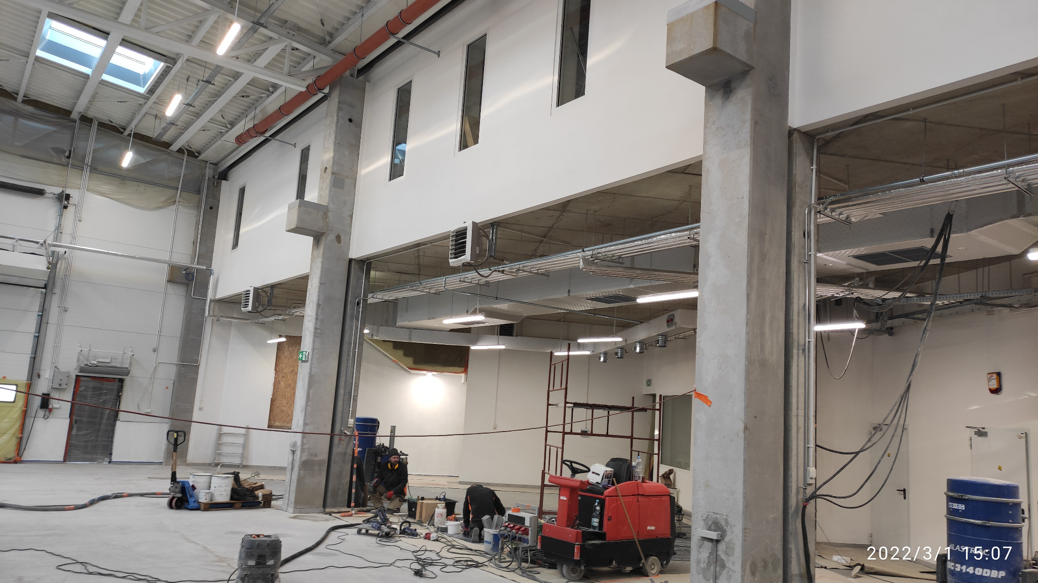 Wnętrze hali produkcyjnej w trakcie budowy: widoczne betonowe słupy, instalacje pod sufitem, okna w górnej części ściany, oraz ekipa budowlana pracująca nad wykończeniem posadzki.
