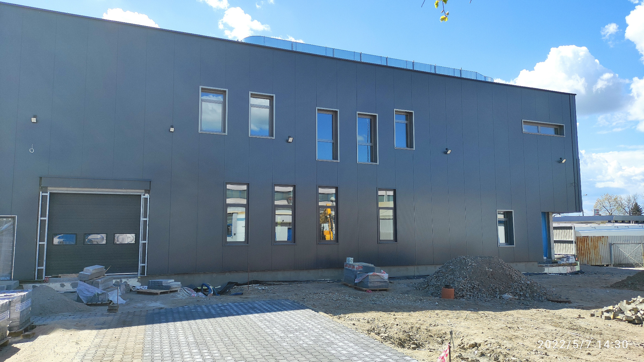 Elewacja frontowa budynku biurowo-produkcyjnego w trakcie budowy, wykonana z szarych płyt warstwowych, widoczne okna z odbiciami, brama garażowa, kostka brukowa i materiały budowlane na placu budowy.