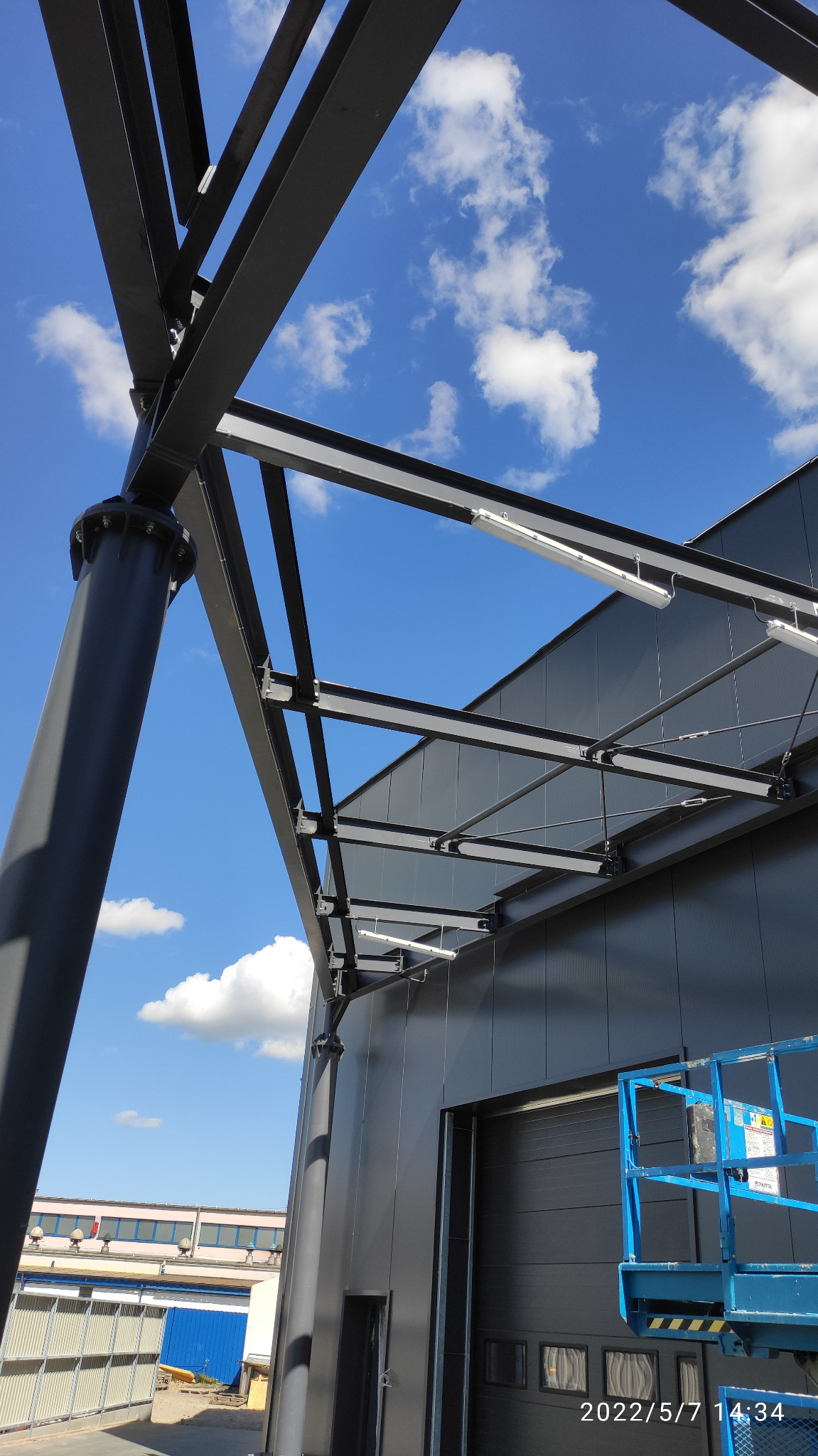 Konstrukcja wiaty magazynowej z czarnych stalowych belek na tle błękitnego nieba z chmurami, widok z dołu, oświetlenie LED pod wiatą, budynek w tle z bramą garażową.