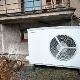 Biała jednostka zewnętrzna pompy ciepła marki Stiebel Eltron ustawiona na betonowych blokach przed budynkiem z widoczną stertą ziemi i gruzu.