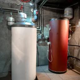 Dwa pionowe zbiorniki akumulacyjne ciepłej wody, biały marki Stiebel Eltron i czerwony marki Kospel, w piwnicy z instalacją hydrauliczną na ścianie z betonu architektonicznego i brudną podłogą...