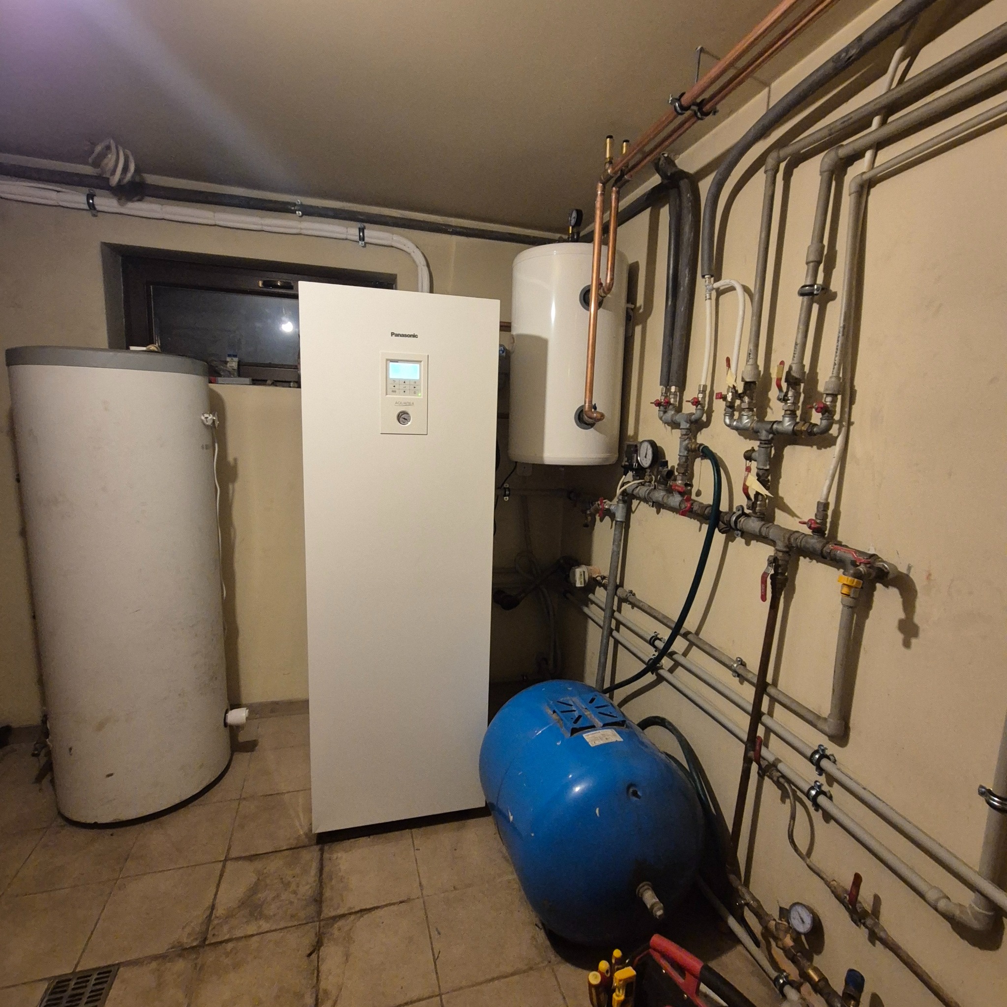 Biała pompa ciepła Panasonic z zbiornikiem, niebieski zbiornik hydroforowy i instalacja rurowa w piwnicy. Widoczne manometry i zawory.