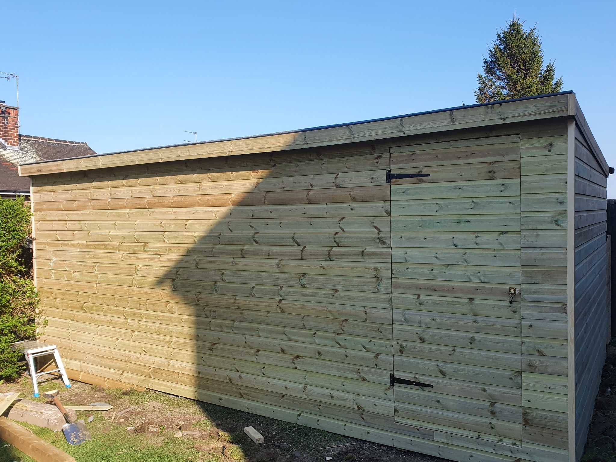 Świeżo postawiony drewniany budynek gospodarczy z pojedynczymi drzwiami, wykonany z impregnowanych desek, widoczna drabinka, łopata i nierówny teren wokół.