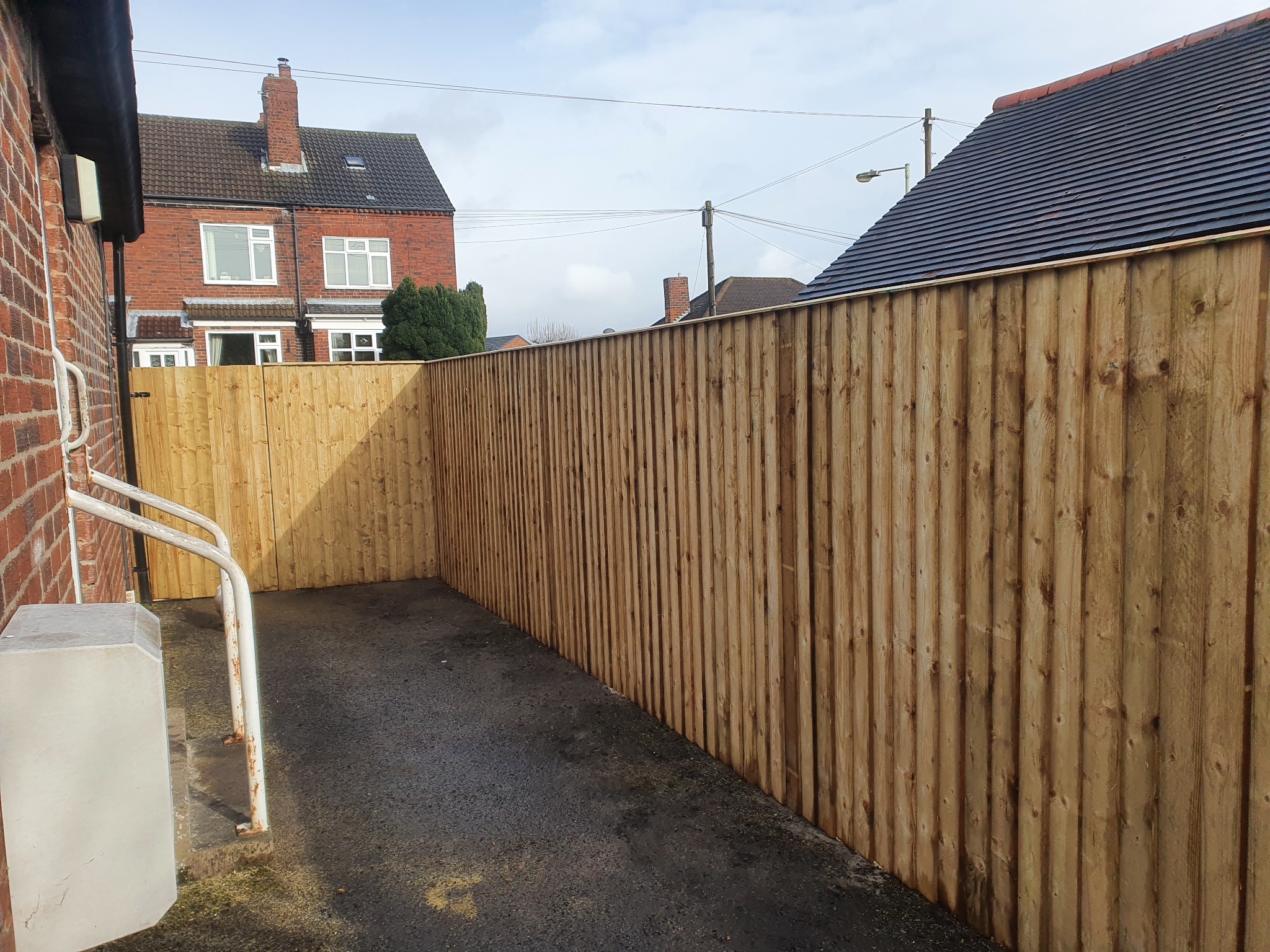 Nowo postawione ogrodzenie z jasnego drewna, pionowe deski, widok z bliska, na tle budynków mieszkalnych i pochmurnego nieba.