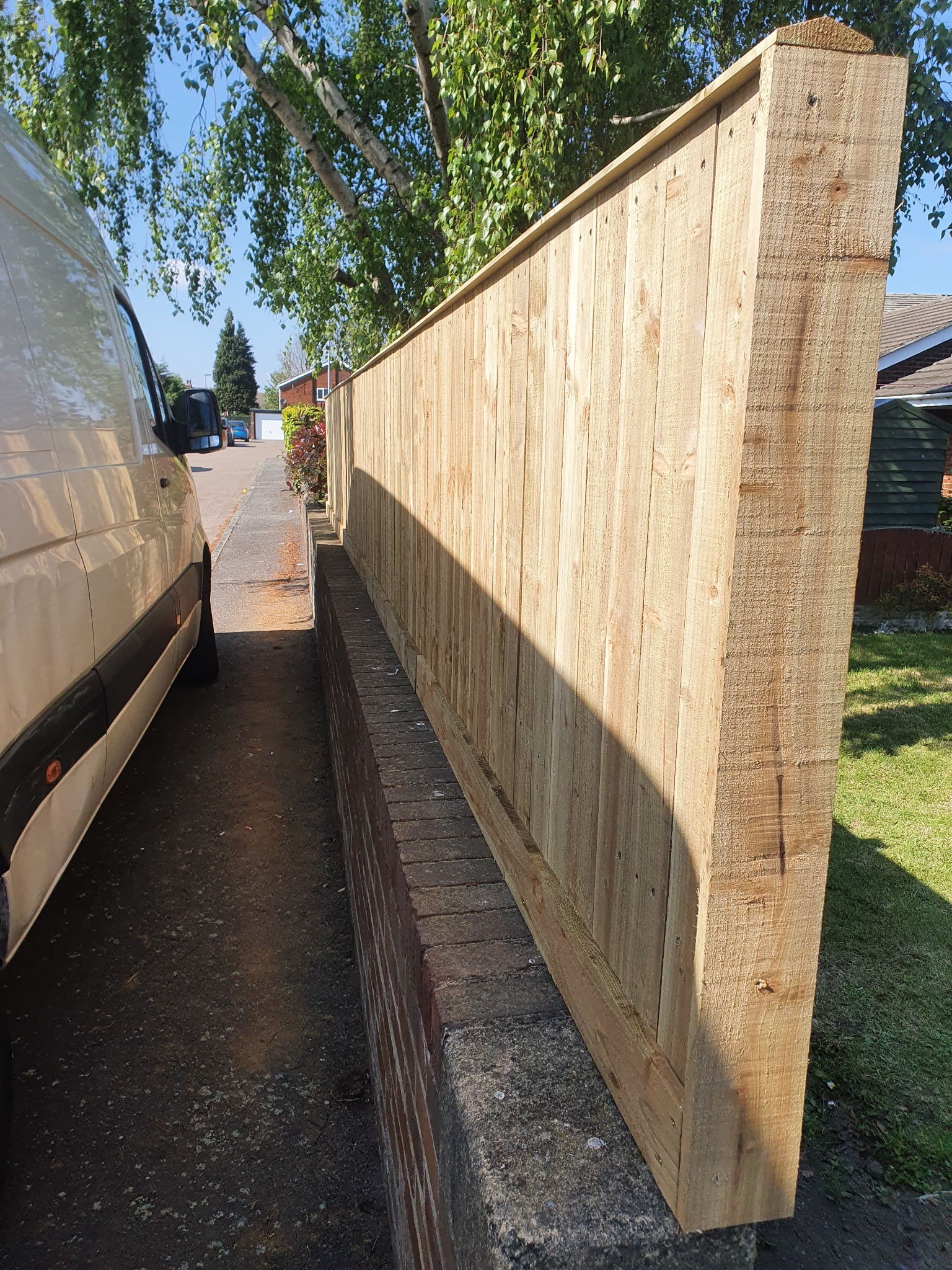 Nowe ogrodzenie z jasnego drewna, widoczne z boku, z zaparkowanym białym vanem obok, na tle zielonego trawnika i domów.