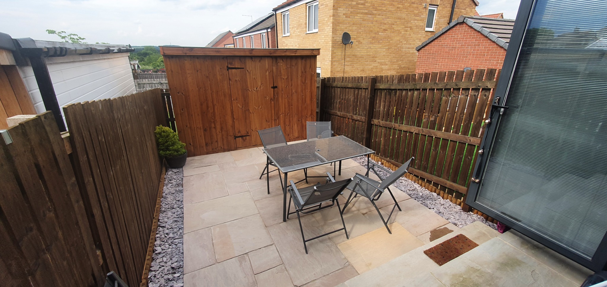 Nowoczesny taras z jasnych płyt, otoczony drewnianym ogrodzeniem i szarą żwirową rabatą, z widocznym drewnianym schowkiem na narzędzia i zestawem mebli ogrodowych z szklanym blatem.