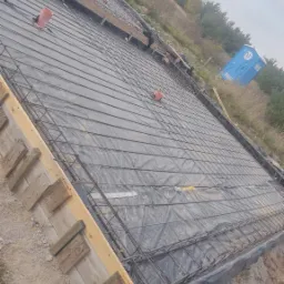 Przygotowanie zbrojenia fundamentu pod dom jednorodzinny, widoczne szalunki z desek, siatka zbrojeniowa na folii, rury wentylacyjne i przenośna toaleta w tle.