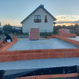 Budowa domu jednorodzinnego: murarz układa pierwszą warstwę cegieł Porotherm na fundamencie, w tle widoczny biały dom z brązowymi zdobieniami okiennymi i zachodzące słońce.