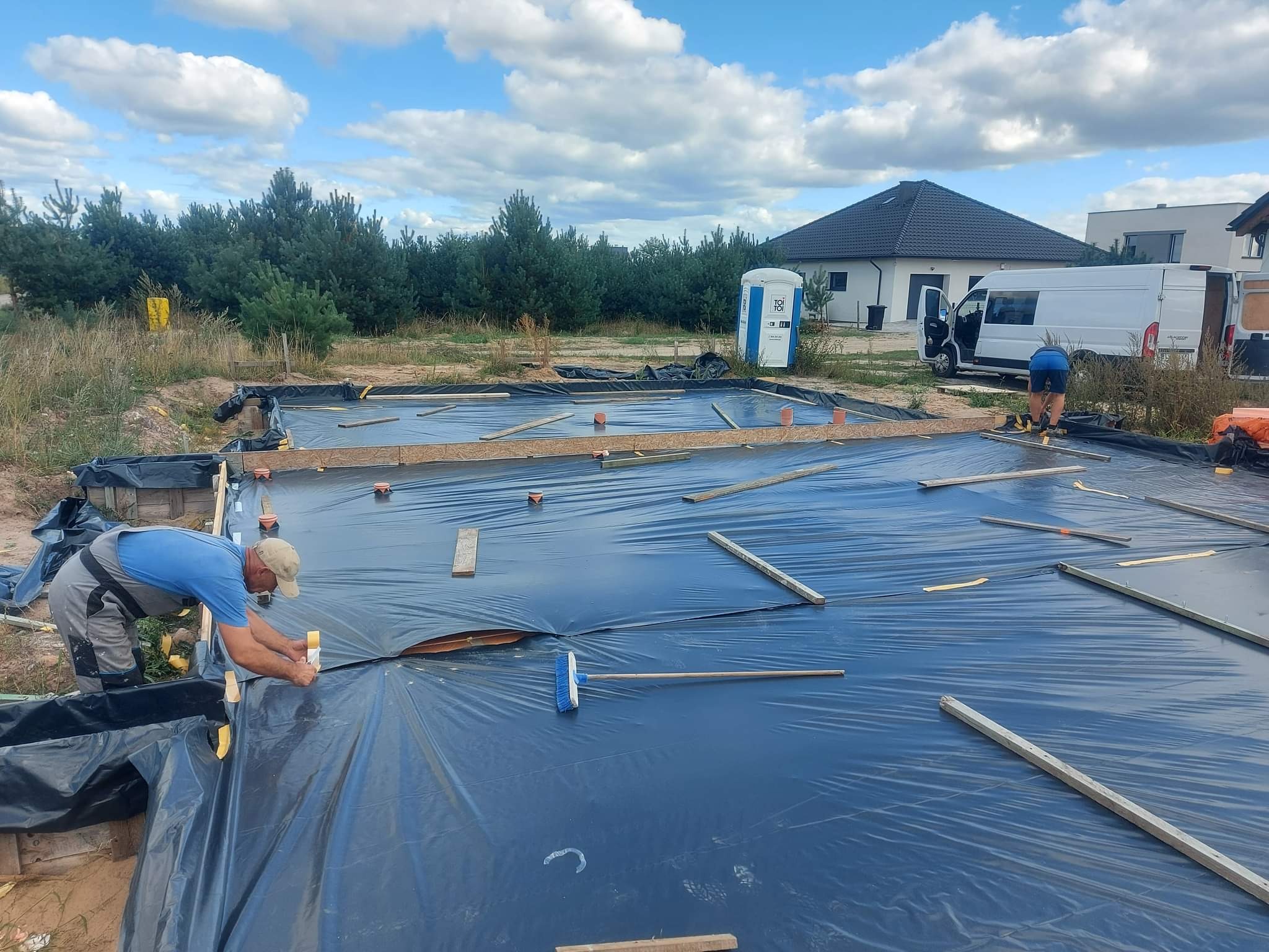 Prace przygotowawcze do wylewki fundamentów: robotnik mocuje folię hydroizolacyjną do drewnianego szalunku, w tle widoczna przenośna toaleta, dom w budowie i zaparkowany biały bus.