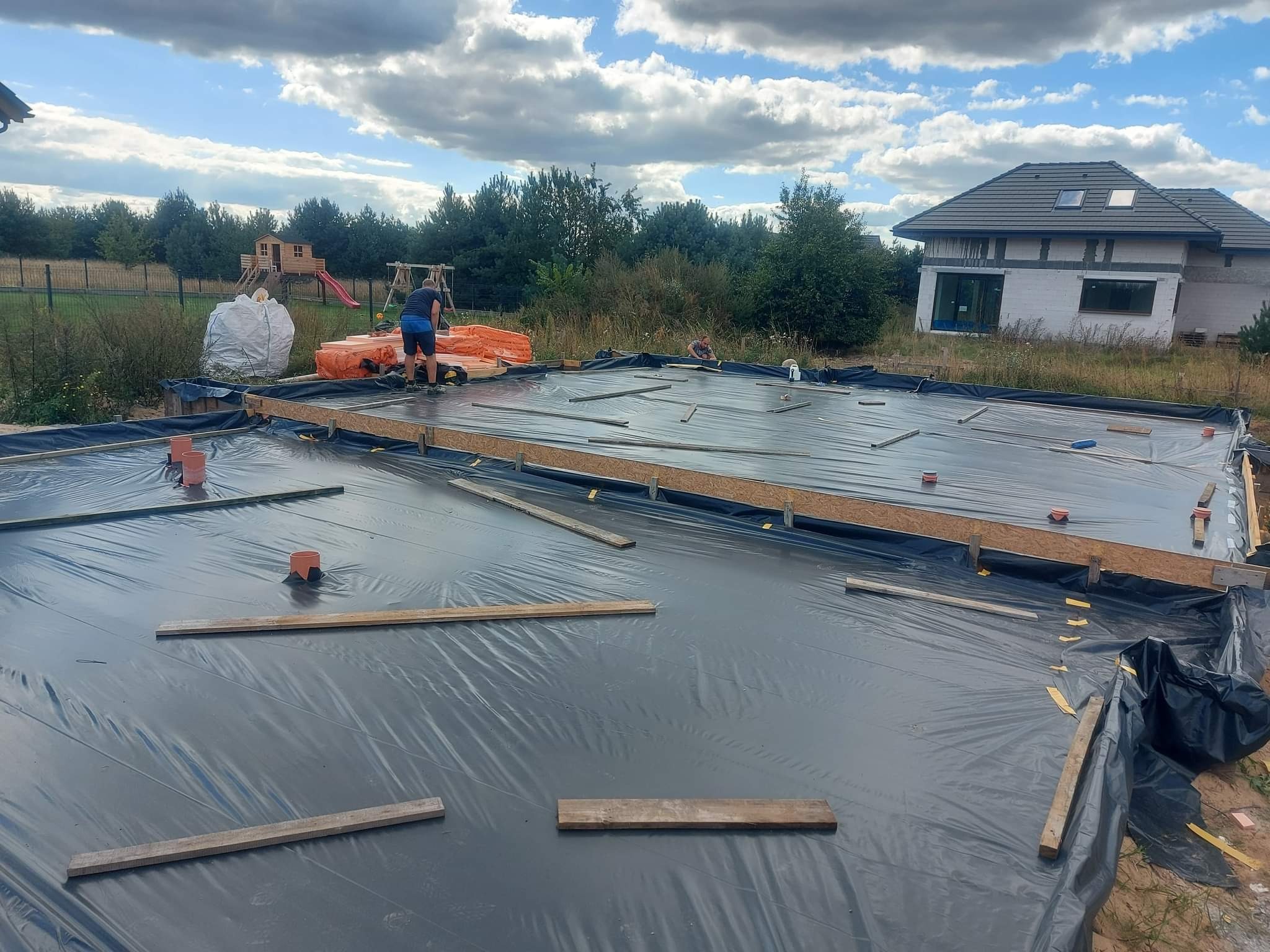 Przygotowanie fundamentów pod budowę domu: rozłożona folia hydroizolacyjna, drewniane szalunki, pracownicy w tle, widoczny budynek w stanie surowym otwartym oraz plac zabaw w oddali.