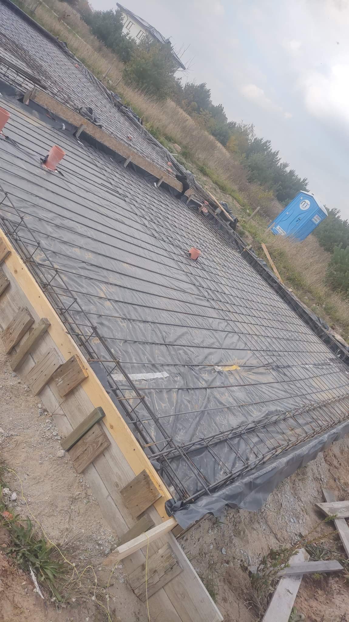 Przygotowanie zbrojenia fundamentu pod dom jednorodzinny, widoczne szalunki z desek, siatka zbrojeniowa na folii, rury wentylacyjne i przenośna toaleta w tle.