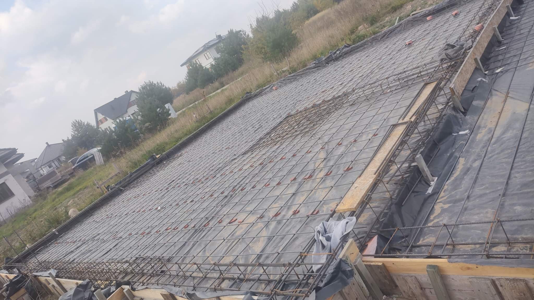 Przygotowanie zbrojenia i szalunku pod płytę fundamentową domu jednorodzinnego, widok z góry na siatkę zbrojeniową ułożoną na folii, drewniane szalunki wzdłuż krawędzi.
