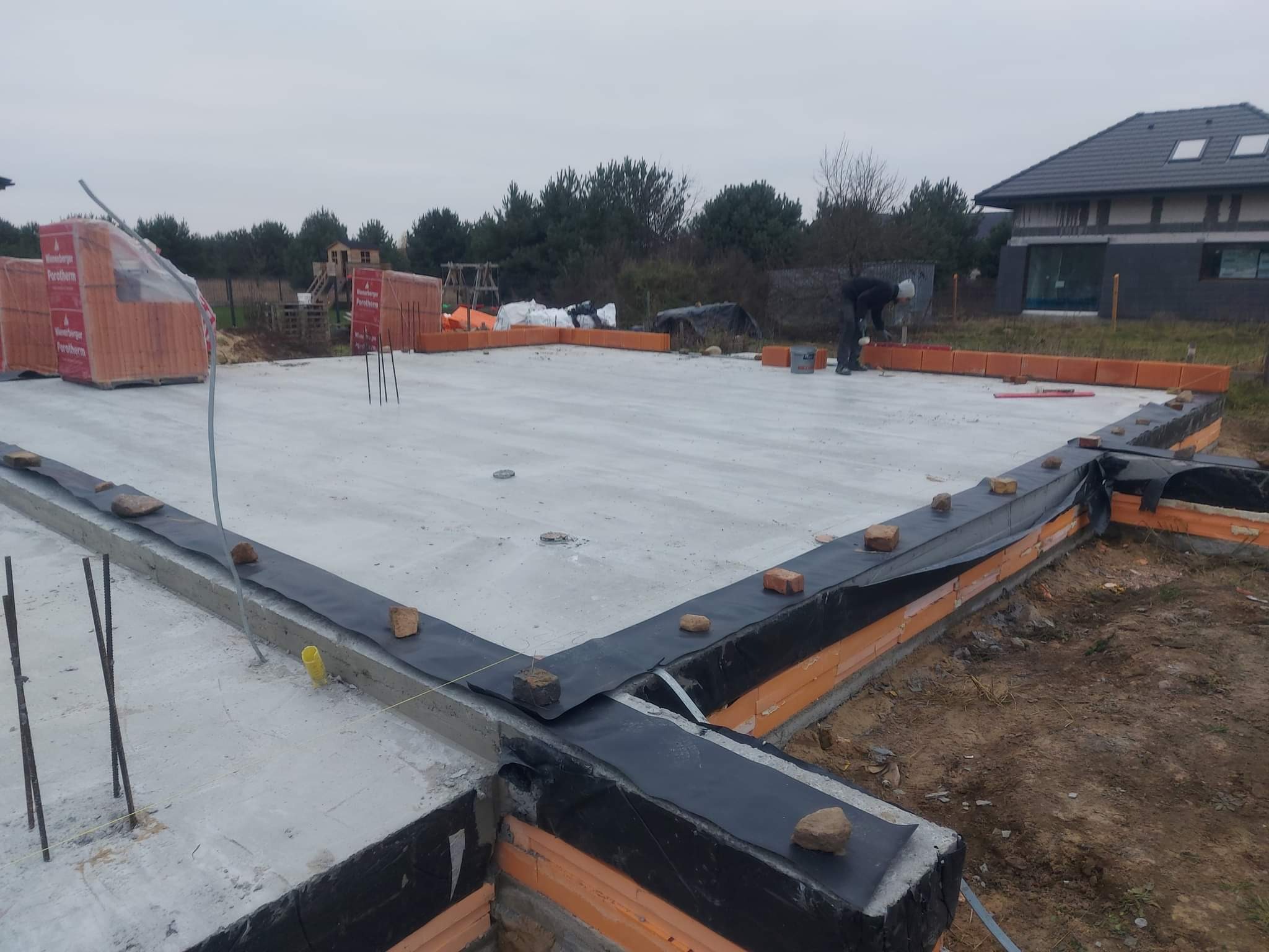 Wylana płyta fundamentowa z izolacją poziomą i rozpoczętym murowaniem pierwszej warstwy ścian z bloczków ceramicznych Porotherm, widoczny pracownik w tle.