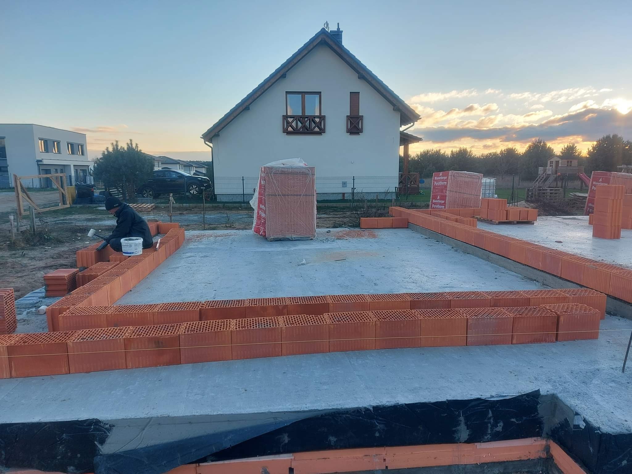 Budowa domu jednorodzinnego: murarz układa pierwszą warstwę cegieł Porotherm na fundamencie, w tle widoczny biały dom z brązowymi zdobieniami okiennymi i zachodzące słońce.