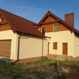 Dwukondygnacyjny dom z garażem, wykończony jasnym tynkiem i drewnianą boazerią pod dachem z brązowej dachówki, widoczny w porannym świetle.