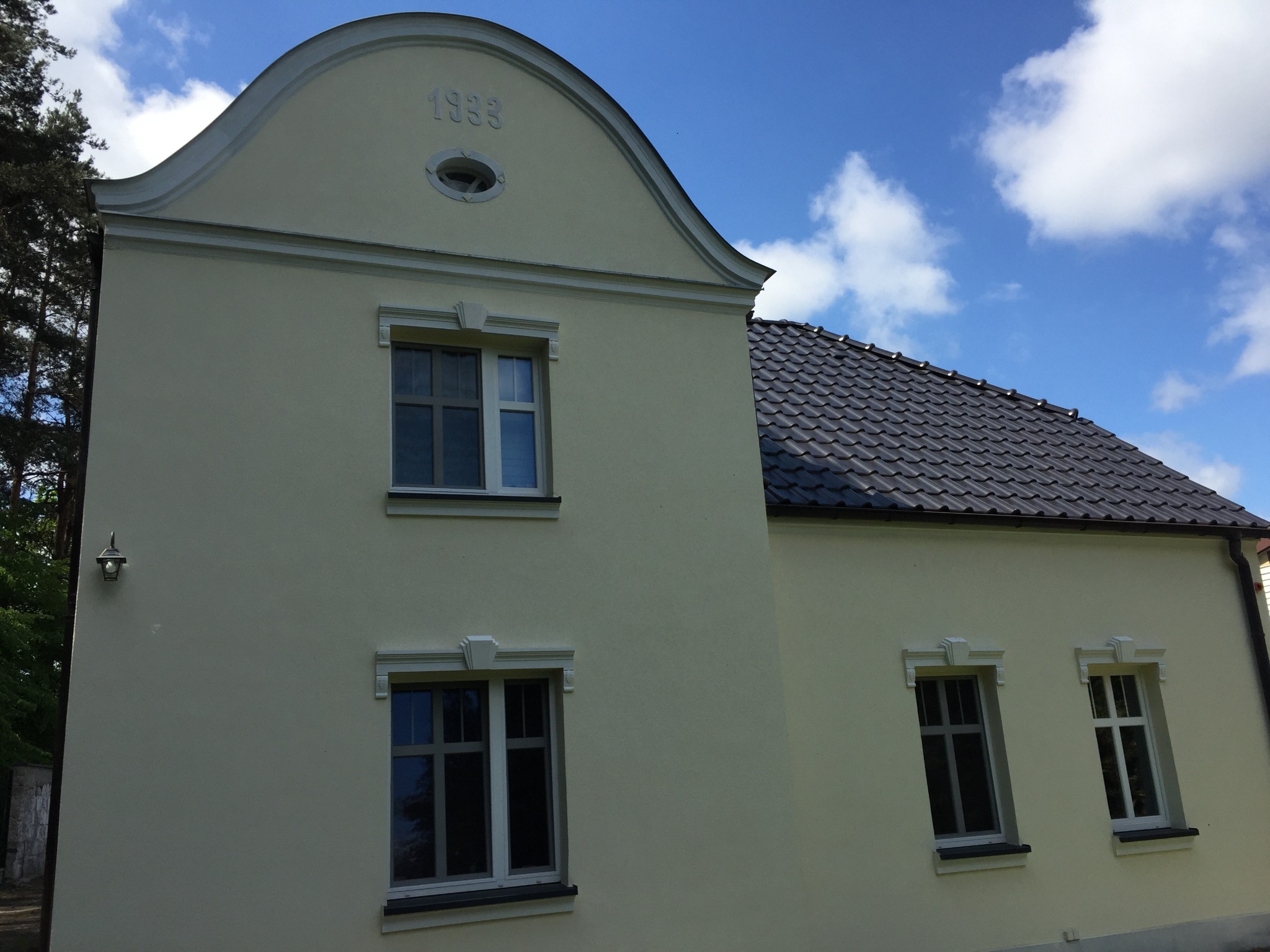 Elewacja odnowionego domu z datą 1933 na szczycie, widoczne okna z dekoracyjnymi obramowaniami i ciemna dachówka
