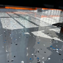 PHO HOME - Świeżo wylana, szara posadzka z kolorowymi płatkami, widoczne refleksy światła i cień konstrukcji nad nią.