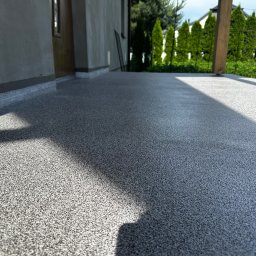 PHO HOME - Szara posadzka żywiczna z drobnym wzorem na tarasie przydomowym, widok z niskiej perspektywy z widocznym fragmentem ściany budynku i słupem pergoli.