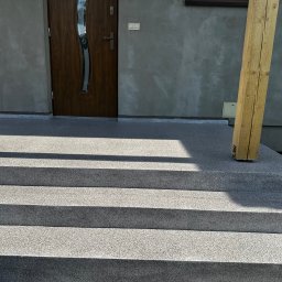 PHO HOME - Schody zewnętrzne z szarą, drobnoziarnistą powierzchnią, prowadzące do drzwi wejściowych domu, z widoczną drewnianą podporą pergoli po prawej stronie.