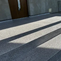 Zewnętrzne schody wejściowe z szarej posadzki o drobnej fakturze, oświetlone mocnym światłem słonecznym tworzącym kontrastowe cienie.