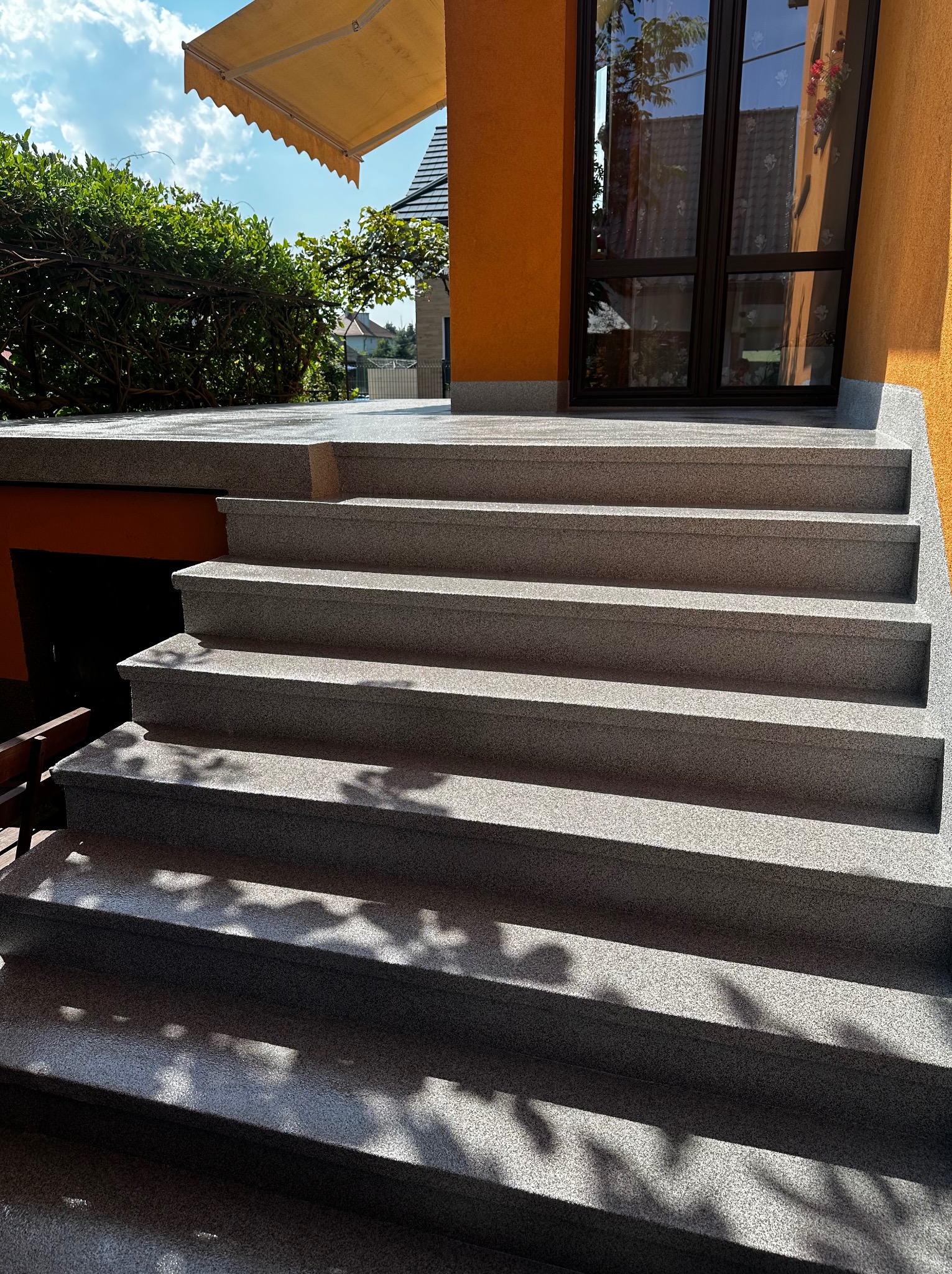 Zewnętrzne schody z szarego kamienia prowadzące do wejścia domu z pomarańczową elewacją, na stopniach widoczne cienie drzew.