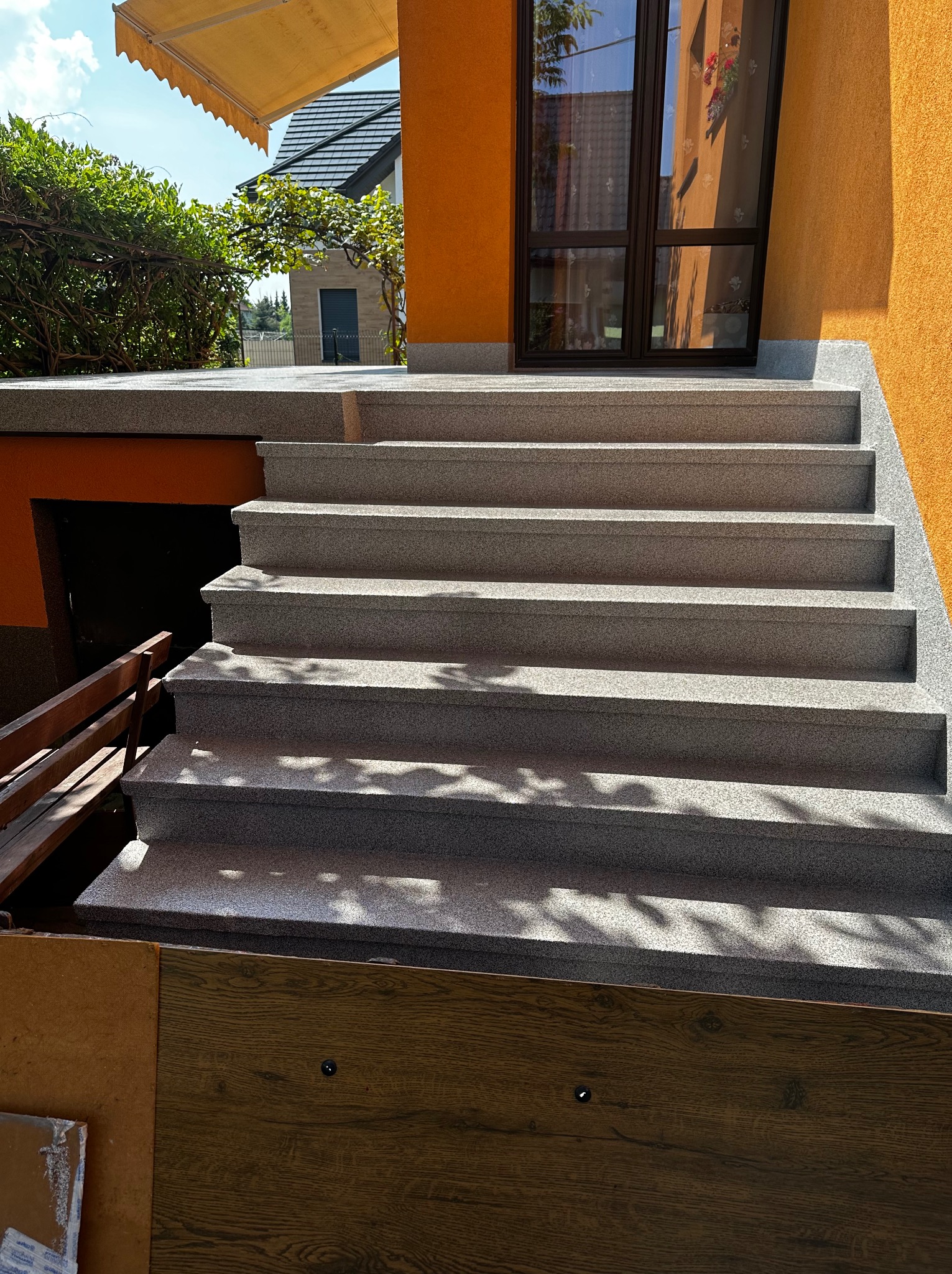 Zewnętrzne schody z szarego kamienia prowadzące do pomarańczowego budynku z ciemnymi oknami, częściowo oświetlone słońcem, z widocznymi cieniami liści