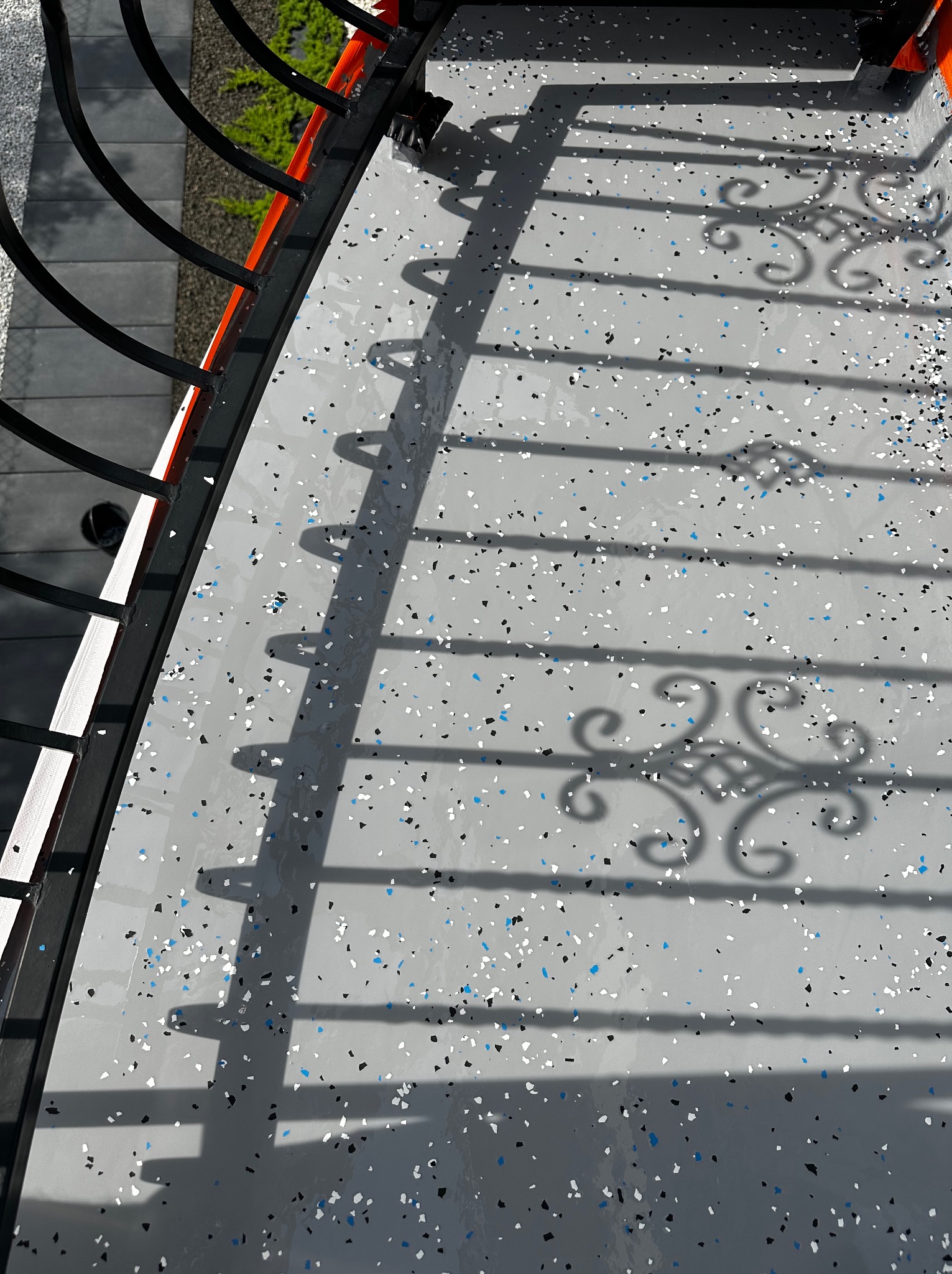 Szara posadzka z kolorowymi drobinkami na balkonie, widoczne cienie balustrady z metalowymi zdobieniami.