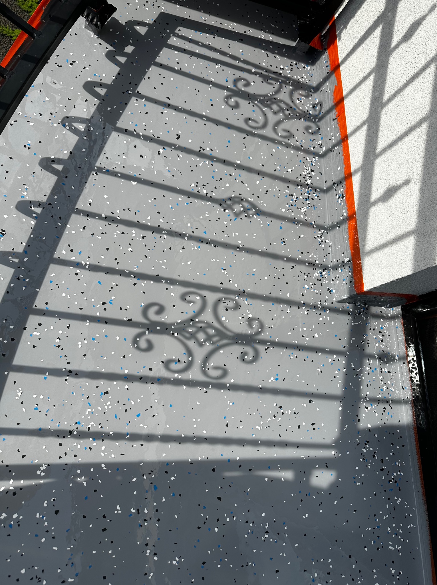 Szara posadzka z kolorowymi płatkami na balkonie, cień ozdobnej balustrady, pomarańczowe wykończenie przy ścianie.