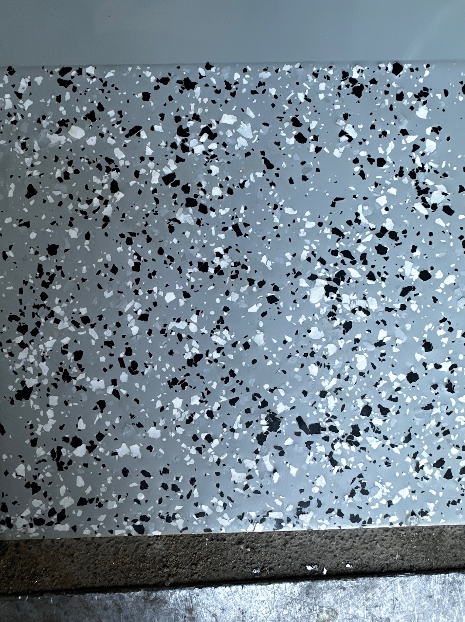 Szara posadzka z czarnymi i białymi wtrąceniami, widoczny fragment krawędzi podłogi.