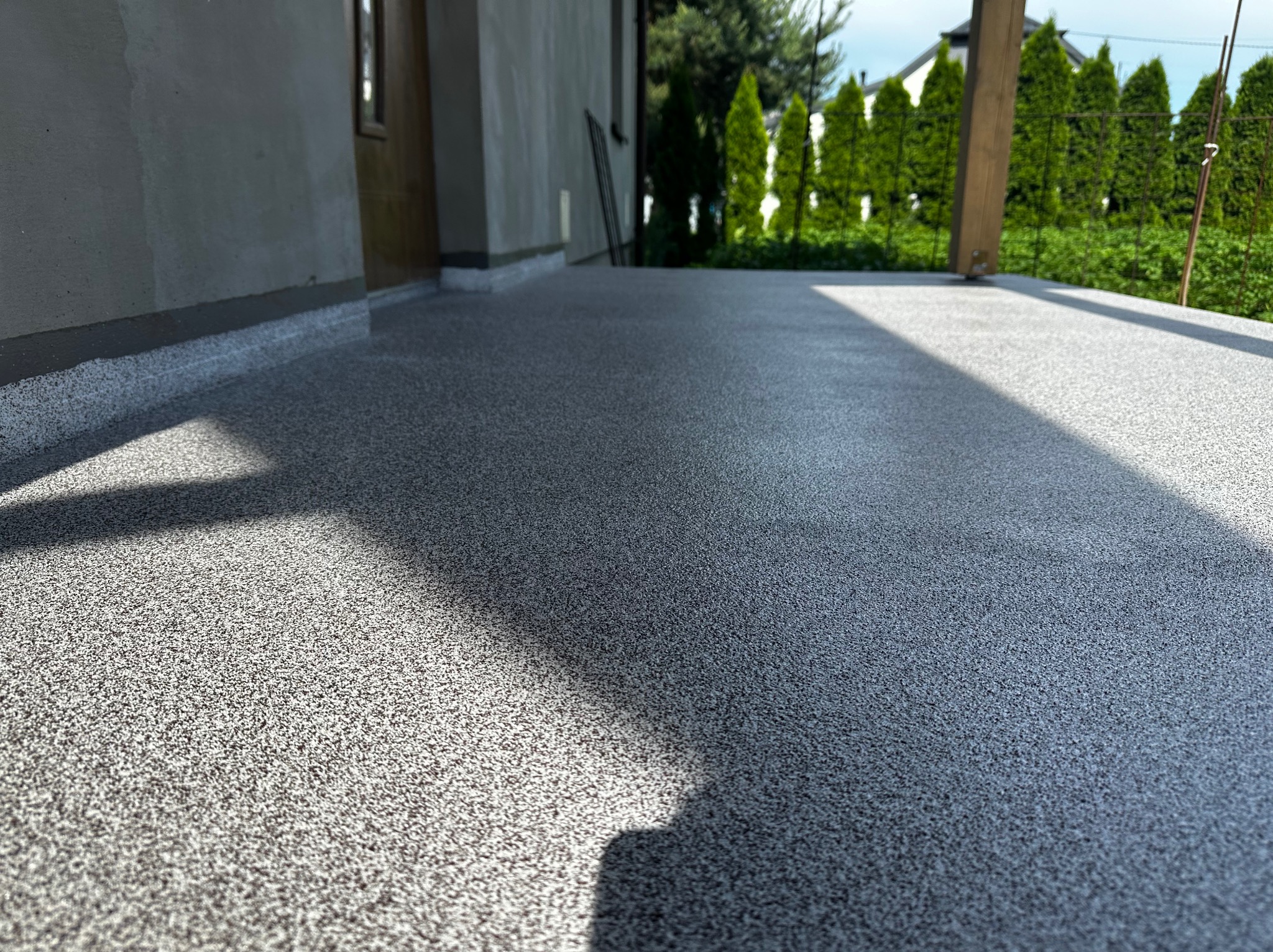 Szara posadzka żywiczna z drobnym wzorem na tarasie przydomowym, widok z niskiej perspektywy z widocznym fragmentem ściany budynku i słupem pergoli.