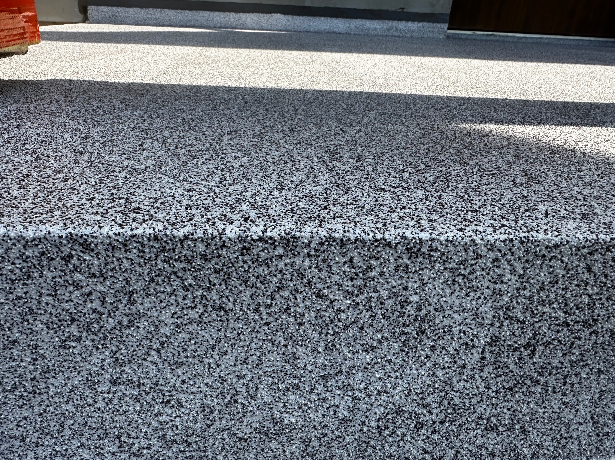 Szara posadzka żywiczna z czarnymi drobinkami, widoczny fragment schodka i cień.