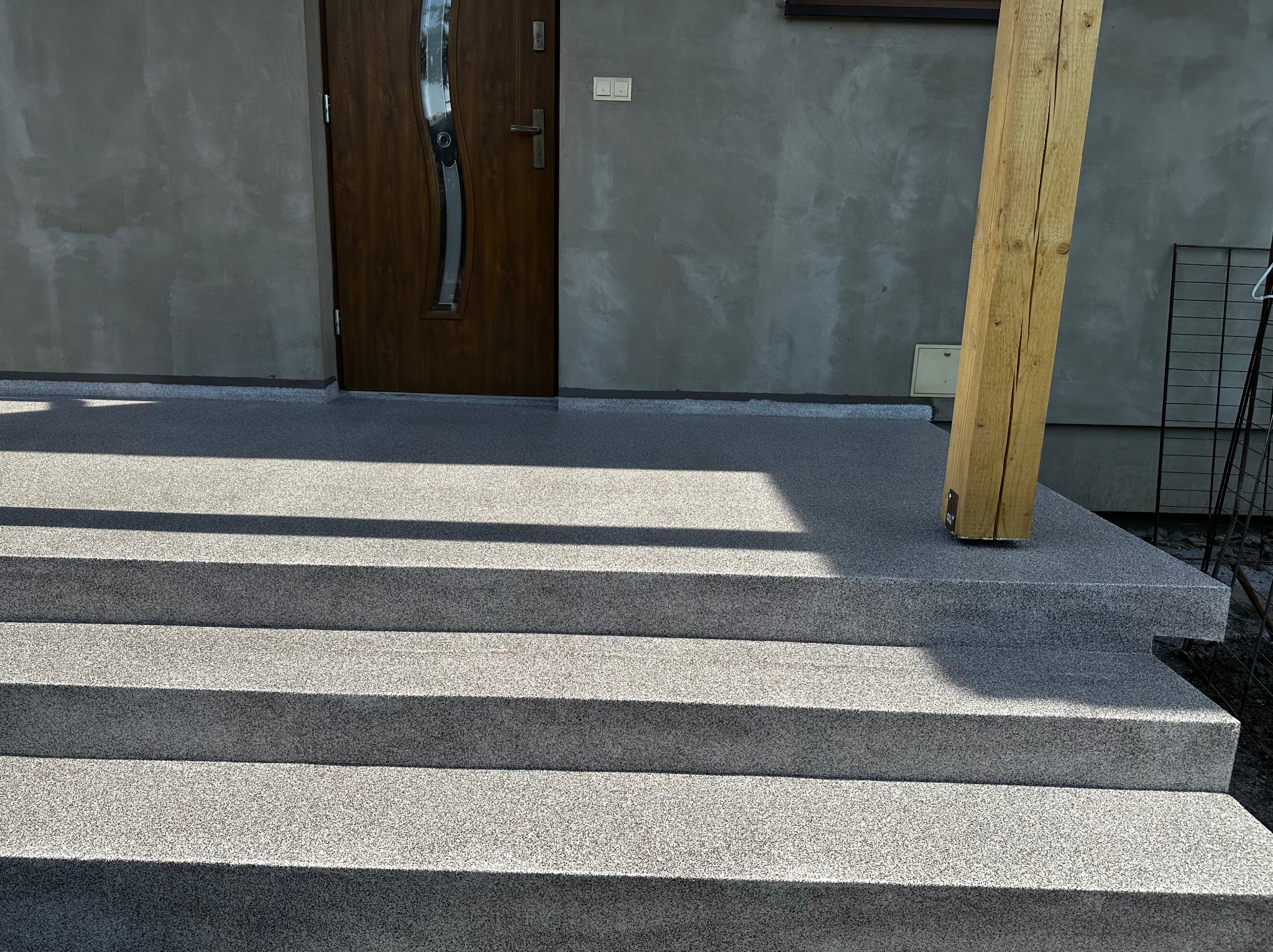 Schody zewnętrzne z szarą, drobnoziarnistą powierzchnią, prowadzące do drzwi wejściowych domu, z widoczną drewnianą podporą pergoli po prawej stronie.