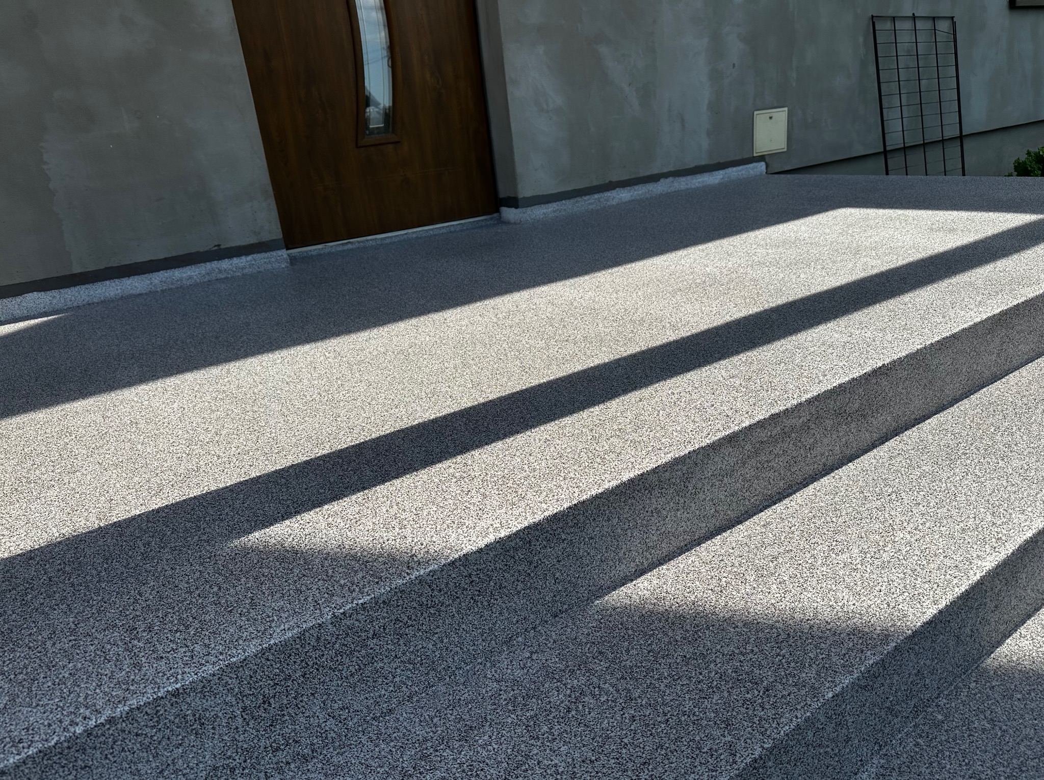 Zewnętrzne schody wejściowe z szarej posadzki o drobnej fakturze, oświetlone mocnym światłem słonecznym tworzącym kontrastowe cienie.