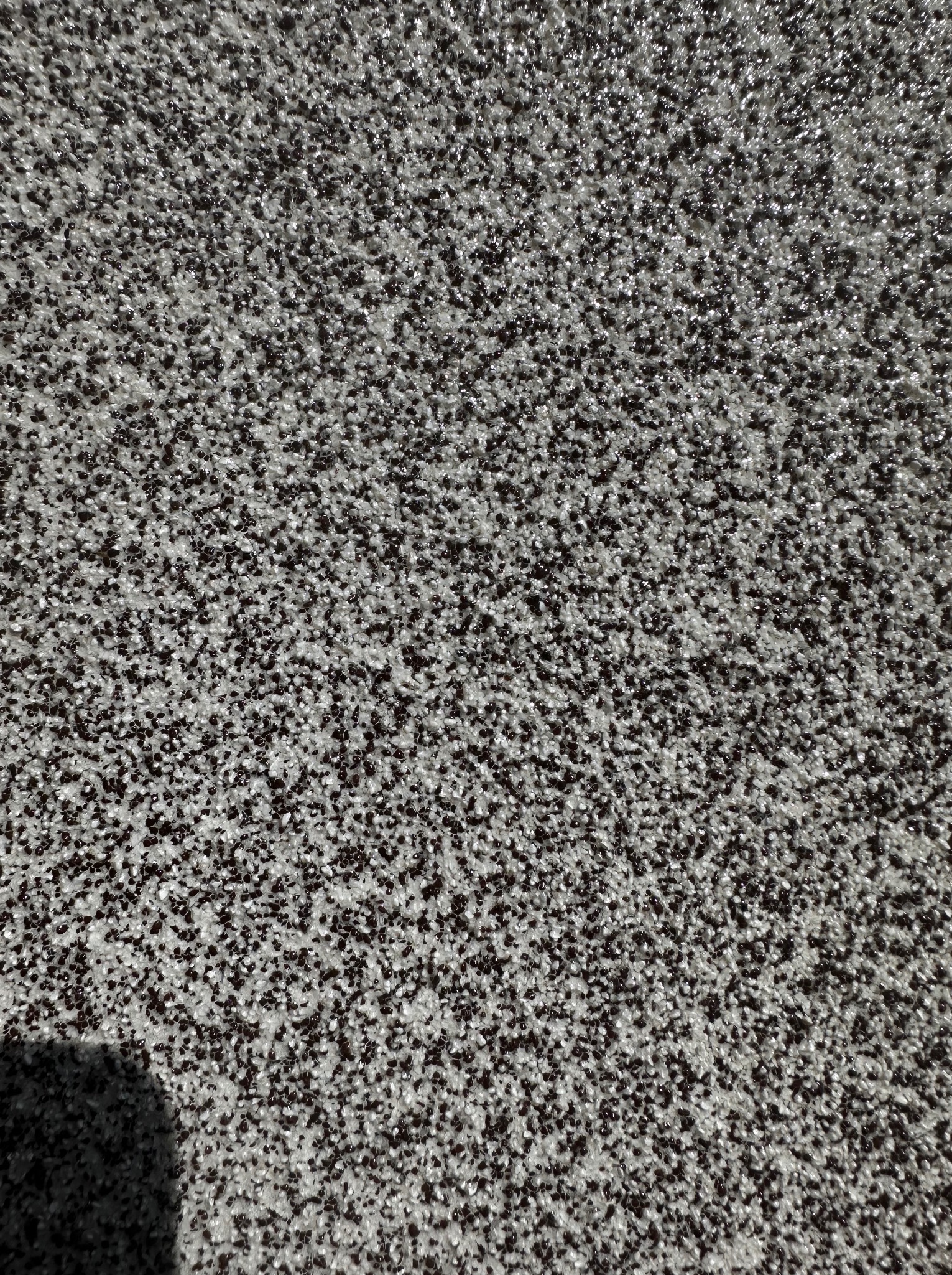 Zbliżenie na szarą posadzkę z drobnymi, ciemnymi granulkami, tworzącymi chropowatą teksturę, widoczny cień w lewym dolnym rogu.